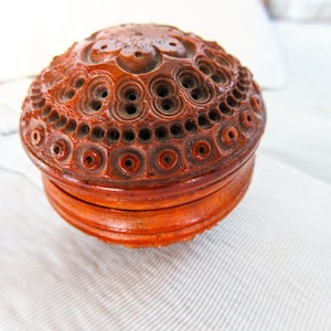 May include: A round, reddish-brown lidded container featuring intricate, carved geometric patterns across its surface. The lid displays a central floral motif surrounded by concentric circles and small, recessed circular indentations.