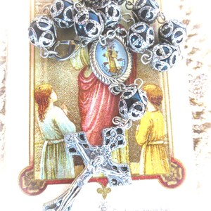 Puede incluir: Un rosario con cuentas negras y plateadas, una cruz plateada y un peque&ntilde;o medall&oacute;n religioso. Las cuentas tienen un patr&oacute;n plateado decorativo. El fondo presenta una imagen religiosa con texto.
