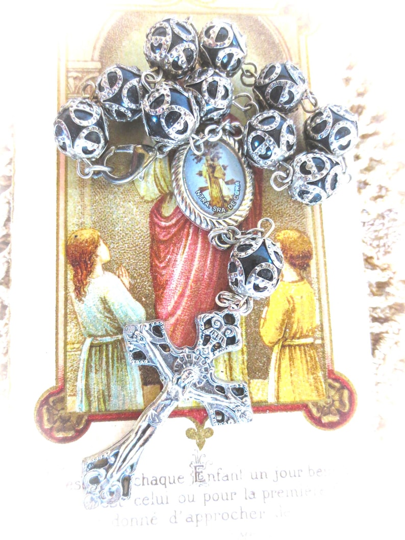 Puede incluir: Un rosario con cuentas negras y plateadas, una cruz plateada y un peque&ntilde;o medall&oacute;n religioso. Las cuentas tienen un patr&oacute;n plateado decorativo. El fondo presenta una imagen religiosa con texto.