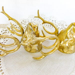 May include: Two gold-colored metal deer head figurines with prominent antlers. Each deer head is sculpted with detailed facial features and textured fur, facing upward in a fixed position. The metallic surface has a polished, reflective finish.