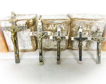May include: Five antique crucifixes with detailed bronze figures of Jesus. The crosses have black and silver accents, some with skull and crossbones. The background features aged, textured blocks with faded text.