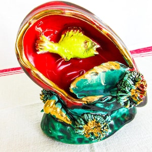 May include: A ceramic shell-shaped bowl featuring a glossy red interior with a bright yellow fish relief. The exterior is textured with green and black tones, accented by gold-painted sea anemones and small shell details along the base.