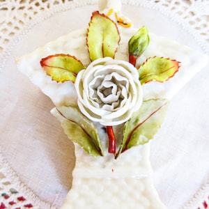 May include: A white porcelain figurine of a single white rose with green leaves and a red stem. The rose is in a white porcelain vase with a textured surface.