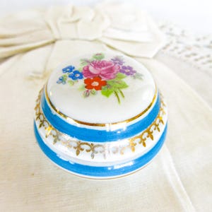 May include: A small, round, porcelain trinket box with a white lid decorated with a floral bouquet of pink, red, blue, and purple flowers. The box has blue stripes with gold accents and a gold rim.