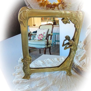 May include: A gold-toned ornate mirror with a decorative floral design and a figure of a woman in the frame. The mirror is sitting on a white crocheted doily.