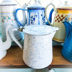 May include: A collection of vintage enamelware teapots in various colors and patterns. The teapots include white with blue speckles, floral designs, and solid blue with daisy accents. They are arranged on a wooden shelf.