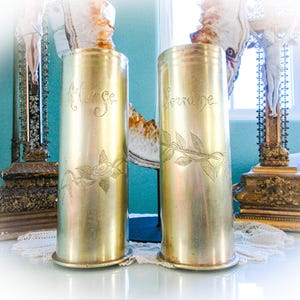 May include: Two antique brass vases with engraved floral designs and the names "Marse" and "Lorraine". The vases are cylindrical and sit on a white surface, with decorative objects in the background.