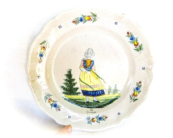 Puede incluir: Plato de cerámica vintage con borde festoneado, con una escena pintada a mano de una mujer con vestimenta tradicional. El plato tiene diseños florales y geométricos en el borde en azul, amarillo y rojo. La mujer lleva un vestido amarillo y sostiene un objeto verde.