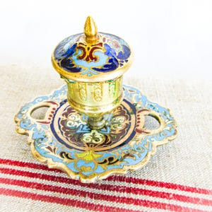 May include: A gold-toned metal inkwell featuring a square base with two handles and a removable lid. The surface is decorated with intricate enamel patterns in shades of light blue, dark blue, red, and green, creating a detailed floral motif.