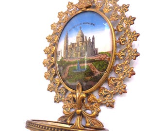 May include: A wall-mounted holy water font featuring an ornate gold-toned metal frame with a circular center depicting the Sacré-Cœur Basilica in Montmartre. The piece includes a small, shallow, light-colored basin at the bottom for holding water.
