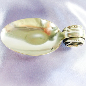 May include: A silver spoon ring with a detailed handle. The spoon bowl is oval-shaped and polished. The ring is made from a repurposed spoon, showcasing a unique design. The ring is set against a soft, lavender background.