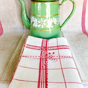 May include: A stack of folded off-white linen towels featuring red checkered patterns and red striped borders, tied with red and white twine. The towels are embroidered with the letters M.P. A green enameled metal coffee pot sits behind them.