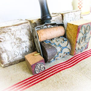 May include: A collection of vintage printing tools including a metal roller with a wooden handle, a small square wooden stamp with a metal monogram, and three large rectangular wooden blocks with white paint and the embossed text SAVON DE LISIEUX.