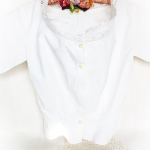 May include: A white cotton blouse with a lace trim and button closure. The blouse is hanging on a wooden hanger with a crocheted flower decoration.
