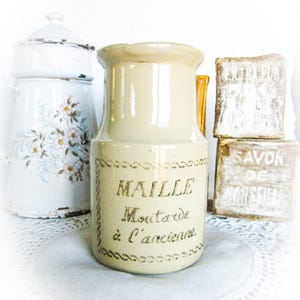 May include: A vintage-style mustard jar with the text "MAILLE Moutarde à l'ancienne" in black script. A white floral enamelware pot and two bars of soap are also visible. The items are arranged on a white lace doily.