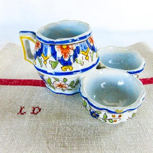 May include: A vintage ceramic creamer and two small bowls. The creamer has a handle and is decorated with blue, orange, and white floral patterns. The bowls and creamer sit on a linen cloth with a red stripe.