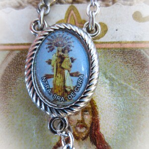 Puede incluir: Colgante religioso plateado con una imagen ovalada azul y dorada de una figura religiosa. El colgante est&aacute; sujeto a una cadena plateada. La imagen est&aacute; rodeada por un dise&ntilde;o de cuerda trenzada.