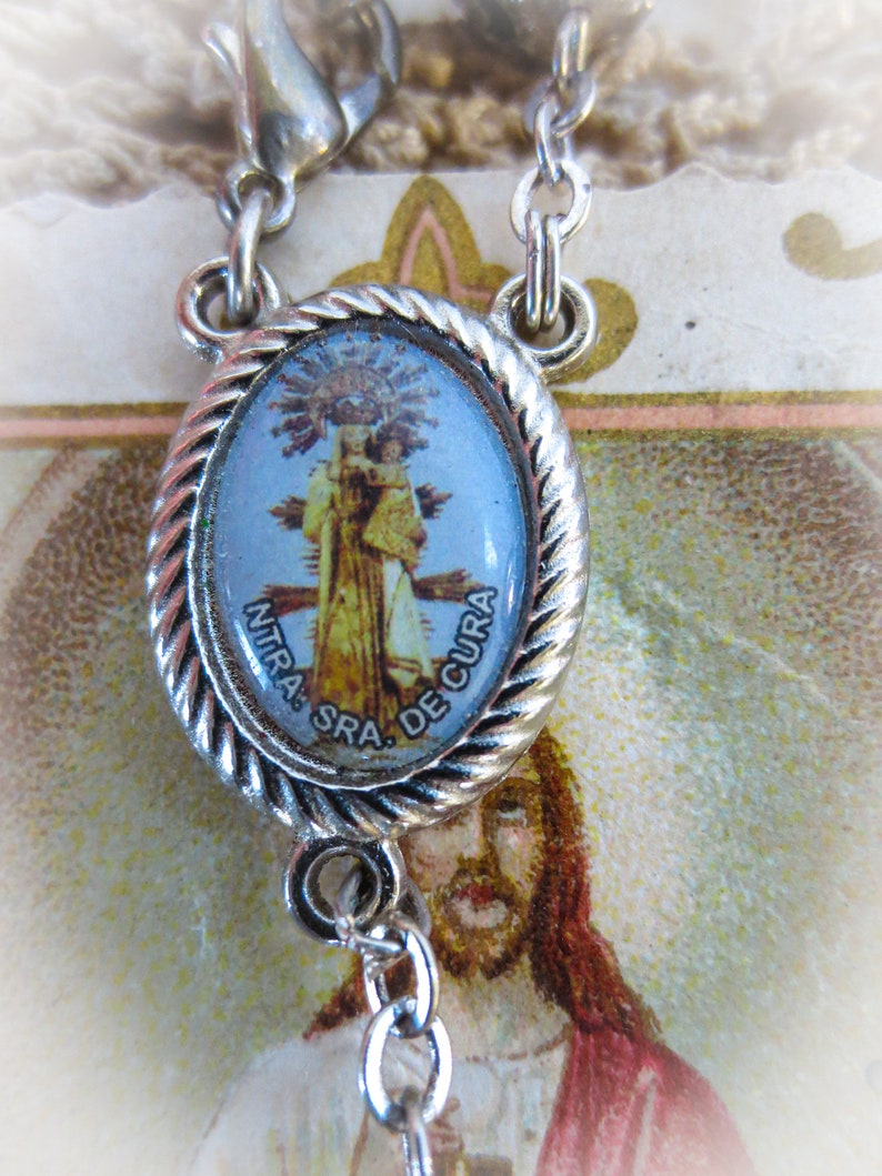 Puede incluir: Colgante religioso plateado con una imagen ovalada azul y dorada de una figura religiosa. El colgante est&aacute; sujeto a una cadena plateada. La imagen est&aacute; rodeada por un dise&ntilde;o de cuerda trenzada.