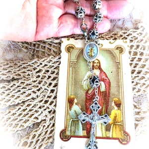 Puede incluir: Un rosario plateado con cuentas negras y plateadas, un colgante de cruz y una tarjeta con una imagen religiosa. La tarjeta presenta una ilustraci&oacute;n de Jes&uacute;s con dos ni&ntilde;os y texto en franc&eacute;s. El rosario se sostiene en una mano.