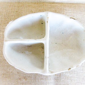 Könnte beinhalten: Eine ovale weiße Keramikschale mit drei getrennten Fächern, die durch interne Trennwände unterteilt sind. Die Oberfläche hat ein glänzendes Finish mit sichtbaren kleinen Unvollkommenheiten und dunklen Punkten in den Innenbereichen.