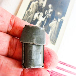 May include: A small, weathered, rectangular metal object with a hinged top flap. The item features a dull, grey, oxidized finish with visible signs of wear and texture across its surface. It is constructed from folded sheet metal components.