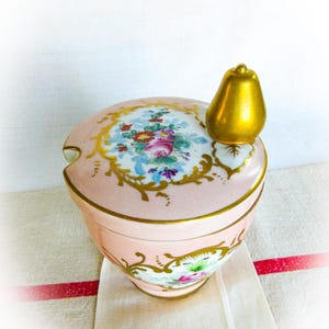 May include: A pink porcelain sugar bowl with a lid decorated with floral patterns and gold trim. The lid has a gold pear-shaped handle. The bowl sits on a white cloth with a red stripe.
