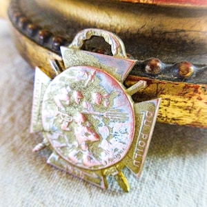 May include: Vintage metal medal with a detailed relief design. The medal features a central circular emblem with figures and text, surrounded by a cross-shaped frame. The colors are gold, bronze, and red.