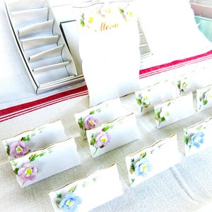 May include: A collection of white porcelain place card holders with gold trim, each adorned with a 3D floral design in shades of pink, blue, and yellow. A matching menu card is also present, along with a silver box.