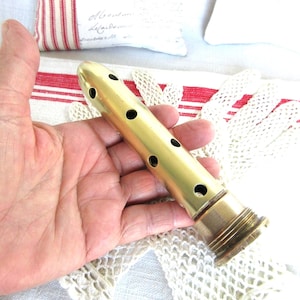 May include: A brass musical instrument with a cylindrical body and several circular holes. The instrument is held in a hand, and the background includes a white and red striped fabric and a white lace doily.
