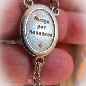 Puede incluir: Primer plano de un colgante de rosario plateado con el texto grabado "Ruega por nosotros". El colgante ovalado est&aacute; conectado a una cadena, con una cuenta decorativa al final. El fondo est&aacute; borroso.