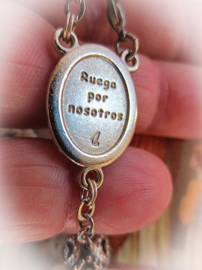 Puede incluir: Primer plano de un colgante de rosario plateado con el texto grabado "Ruega por nosotros". El colgante ovalado est&aacute; conectado a una cadena, con una cuenta decorativa al final. El fondo est&aacute; borroso.