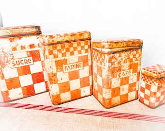 May include: A set of four rectangular metal storage canisters featuring a distressed orange and white checkered pattern. Each container has a lid and is labeled with the French words SUCRE, FARINE, CAFE, and POIVRE in a rectangular frame.