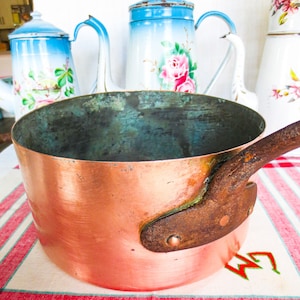 Könnte beinhalten: Ein Kupfertopf mit einem rostigen Griff steht auf einer rot-weiß gestreiften Tischdecke. Der Topf ist leer und hat ein abgenutztes, altes Aussehen.