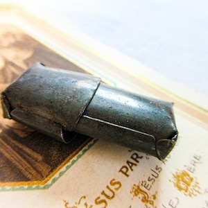 May include: A small, rectangular, dark gray metal object with a folded, overlapping construction. The surface appears weathered with a matte, slightly textured finish, featuring visible seams where the metal has been bent into a compact shape.