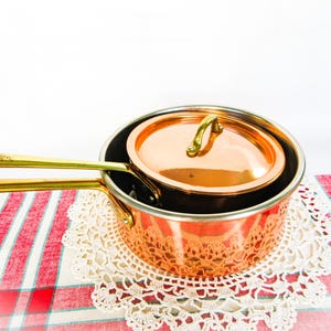 May include: A polished copper saucepan with a matching lid and a long brass handle. The pan features a metallic finish and a sturdy construction, designed for stovetop use. The handle is securely attached to the side of the round copper vessel.
