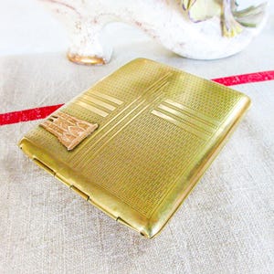 May include: A rectangular, gold-toned cigarette case with a textured surface and a decorative rectangular emblem. The case features etched linear and geometric patterns. The case is resting on a white and beige fabric with a red stripe.