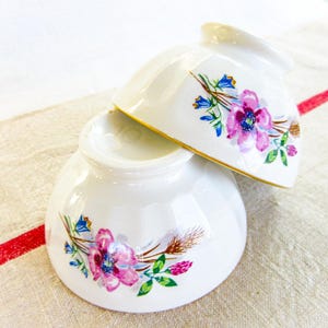 May include: Two white ceramic bowls, one atop the other, with floral designs. The bowls have a gold rim and are decorated with pink, purple, and blue flowers and green leaves. The bowls are on a cream-colored surface.