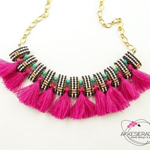 Op de afbeelding: Een zwart-witte kralenketting met turquoise accenten en roze kwastjes. De ketting ligt op een witte achtergrond.