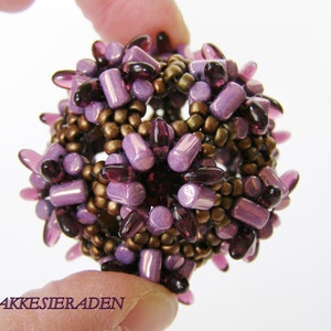 Könnte beinhalten: Eine purpurne und braune Perlenkugel mit geometrischem Muster. Die Perlen sind in einem Muster aus Spitzen angeordnet.