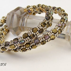 May include: Two gold and silver beaded bracelets with iridescent green and purple beads. The bracelets are intertwined and resting on a clear glass object.