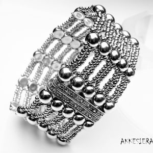 Op de afbeelding: Een zilveren armband met meerdere rijen kralen en een decoratieve sluiting. De armband is gemaakt van metaal en heeft een glanzende afwerking.