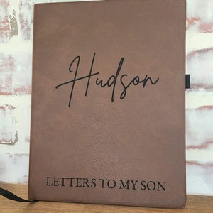 Pu&ograve; includere: Un diario in pelle marrone con il nome "Hudson" in corsivo e il testo "LETTERS TO MY SON" stampato sotto. Il diario ha un segnalibro a nastro nero.
