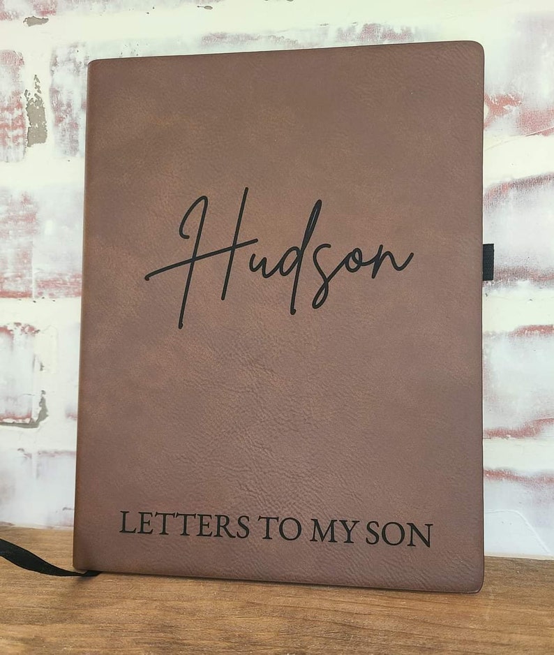 Pu&ograve; includere: Un diario in pelle marrone con il nome "Hudson" in corsivo e il testo "LETTERS TO MY SON" stampato sotto. Il diario ha un segnalibro a nastro nero.