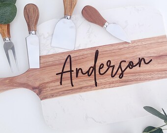 May include: A white and brown wooden cheese board with a handle. The board has a marble-like design on one side and the name "Anderson" engraved on the other side. The board is accompanied by four cheese knives.