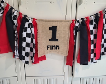 May include: A burlap banner with the number "1" and the name "Finn" in black lettering. The banner is decorated with red, black, and white checkered fabric.