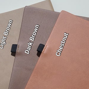 Pu&ograve; includere: Primo piano di tre quaderni in pelle nei colori marrone chiaro, marrone scuro e castagno. Ogni quaderno ha un elastico nero e il nome del colore stampato sulla copertina.