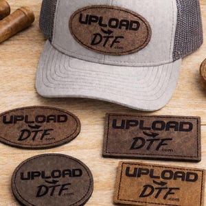 May include: A light gray and mesh trucker hat with a brown leather patch that reads "UPLOAD DTF". Also shown are leather patches, scissors, and a measuring tape on a wooden surface.
