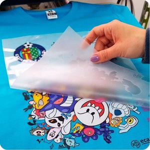 Pode incluir: Uma folha de pl&aacute;stico transparente est&aacute; sendo descascada de uma camiseta azul com uma estampa gr&aacute;fica de desenho animado colorida. O gr&aacute;fico apresenta uma variedade de personagens, incluindo um fantasma, uma caveira e um cogumelo.