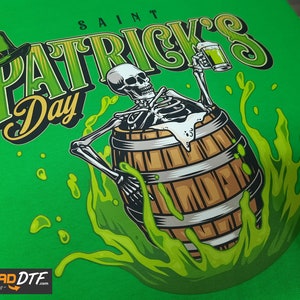 Pode incluir: Uma camiseta verde com um gr&aacute;fico de um esqueleto segurando uma caneca de cerveja e sentado em um barril de l&iacute;quido verde. O texto "SAINT PATRICK'S Day" est&aacute; escrito em uma fonte estilizada acima do esqueleto.