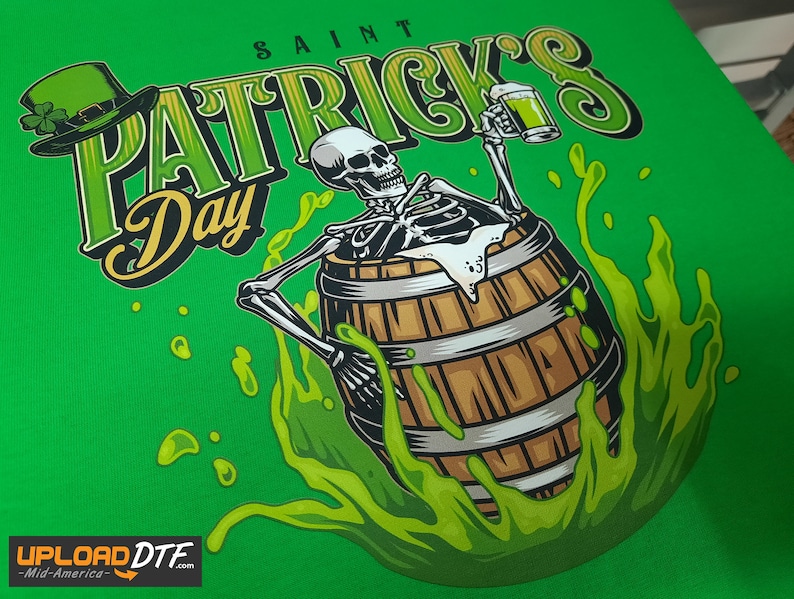Pode incluir: Uma camiseta verde com um gr&aacute;fico de um esqueleto segurando uma caneca de cerveja e sentado em um barril de l&iacute;quido verde. O texto "SAINT PATRICK'S Day" est&aacute; escrito em uma fonte estilizada acima do esqueleto.